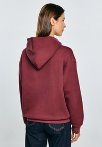 Sudadera marrón burdeos de gran tamaño con un acabado texturizado y un gran bolsillo trasero. Presenta una capucha con cordones y puños acanalados, combinada con vaqueros azules.