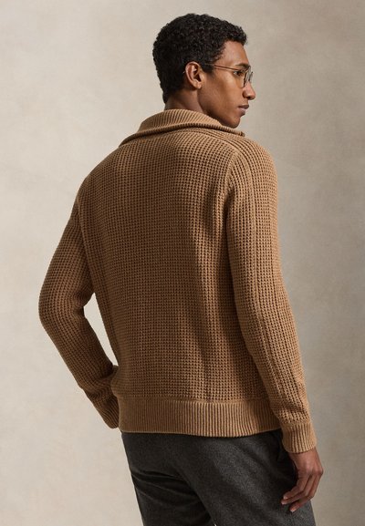 Bărbat cu păr scurt și creț, purtând ochelari și un pulover maro din tricot texturat, cu fața într-o parte, pe un fundal simplu.