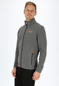 Gray fleece jacket with a high collar, featuring an orange zipper and logo. Includes two zippered side pockets and a fitted design.