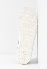 Suola di sneaker bianca con una superficie texture, caratterizzata da una forma curva e un logo impressa sul lato. Il materiale appare gommoso con una finitura liscia.