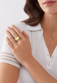 Weißes geripptes Polohemd mit Kragen, ausgestattet mit drei Knöpfen und beigen Streifen an den Ärmeln; trägt einen matten goldenen rechteckigen Ring.