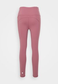 Vista traseira de leggings mauve de cintura alta com detalhes de costura visíveis e um pequeno logótipo refletor próximo ao tornozelo.