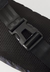 Tissu en maille noire avec une surface texturée, doté d'une boucle en plastique noir pour la fermeture et d'une sangle ajustable. Les motifs sont subtils dans le matériau.