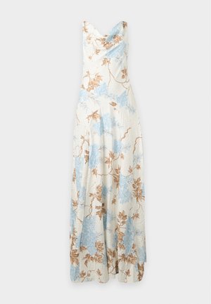 Femme aux longs cheveux foncés portant une robe crème sans manches col bénitier avec imprimé floral bleu et marron, debout les mains dans le dos.