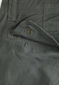 Grauer Stoffbeutel mit sichtbaren Nähten, metallischem Druckknopf und einem zusätzlichen Nietdetail an einer Hose. Einfaches Design, glatte Textur.