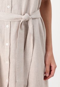 Beige linen dress with a tie belt, featuring a button-down front and a soft, textured fabric. Close-up of hand on the waist.