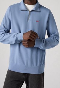 Ljusblå tröja med halvdragkedja, ribbade muddar och Levi's-logotyp på bröstet. Mjuk material, avslappnad passform.