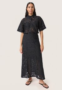 Ensemble en dentelle noire composé d'un haut à manches courtes avec col et d'une longue jupe. Le tissu présente des motifs floraux et une apparence texturée.