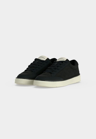 Paire de baskets basses noires avec semelles blanches et lacets noirs, présentée légèrement en angle sur un fond clair uni.