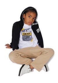 Zwarte zip-up hoodie met logo-accenten, witte graphic tee met gele en grijze tekst, beige broek en witte sneakers met zwarte accenten.