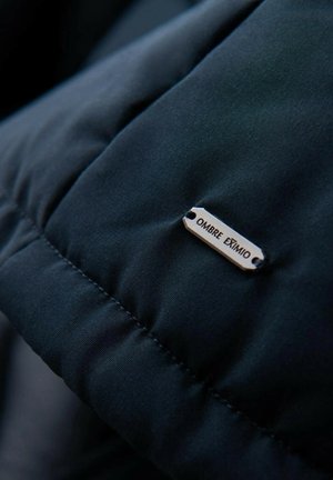 Schwarze Pufferjacke mit gestepptem Muster und einem Metallschild mit der Aufschrift "OMBRÉ EXTIMO", das mit zwei Löchern am Stoff befestigt ist.