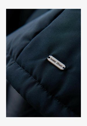 Czarna kurtka puchowa z pikowanym wzorem oraz metalową etykietą oznaczoną "OMBRÉ EXTIMO", przymocowaną do materiału za pomocą dwóch dziurek.