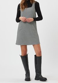 Robe trapèze pied-de-poule noir et blanc avec bretelles réglables, dotée de poches et assortie à des bottes en caoutchouc noires montantes jusqu'aux genoux.