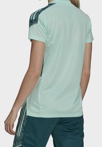 Osoba oblečená ve světle mátově zeleném sportovním tričku s krátkým rukávem, které má tmavě zelené vzorované panely na ramenou a po stranách, doplněné tmavě zelenými kalhotami.