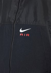 Nike-jacka med svart design, vadderad textur och en vit swoosh-logotyp. "AIR" är tryckt i rött på vänster sida under logotypen.