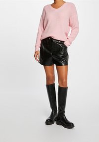 Pull rose clair à col en V avec un tricot texturé, associé à un short en similicuir noir. Bottes noires montant au genou avec une semelle épaisse.