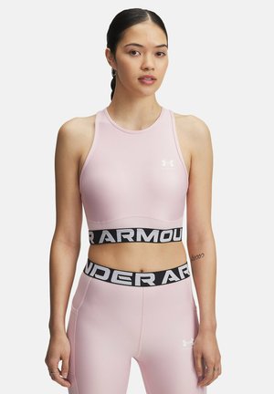 Joven mujer con cabello oscuro vistiendo un sujetador deportivo de color rosa claro de Under Armour y leggings a juego, contra un fondo blanco liso.