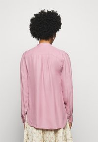 Světle růžová blůza s dlouhým rukávem, se záhybem na zadní straně, knoflíkovými manžetami a jemným, hladkým materiálem. Zakřivený lem, uvolněný střih.