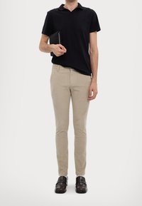 Beżowe spodnie o wąskim kroju, czarna koszulka polo oraz ciemne skórzane mokasyny; mężczyzna trzymający czarny tablet z zamkiem błyskawicznym.