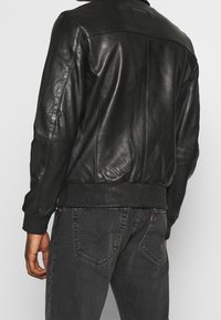 Svart läder bomberjacka med ribbade ärmslut och midjeresår, med en slät textur och minimalistisk design, sedd från sidan.