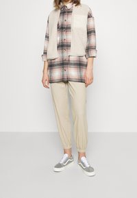 Camicia a scacchi nei toni del rosa e del nero, con un gilet in pile beige. Pantaloni jogger color sabbia e sneakers grigie con lacci bianchi. Vestibilità su misura.