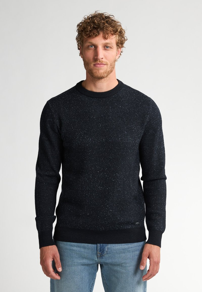 Donkerblauwe gebreide trui met een textuurpatroon, voorzien van een ronde halslijn en geribbelde manchetten. Gecombineerd met lichtblauwe jeans.