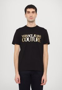 Juodas medvilnės marškinėliai trumpomis rankovėmis, su ryškiu auksiniu užrašų "VERSACE JEANS COUTURE" priekyje. Paprastas, klasikinio kirpimo dizainas.