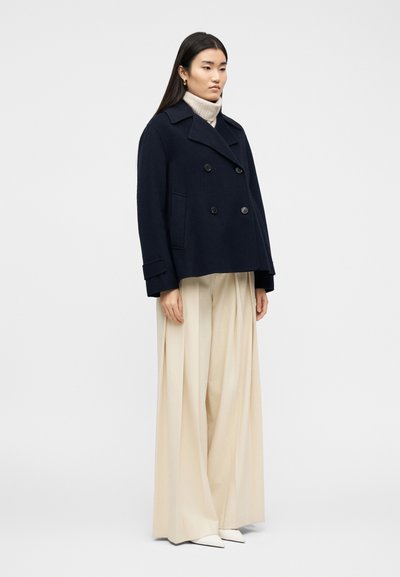 Γυναίκα που φορεί μπλε διπλόμπουτο παλτό τύπου pea coat, μπεζ παντελόνι με φαρδιά μπατζάκια, λευκά μυτερά παπούτσια και ένα μπεζ πουλόβερ με ψηλό λαιμό, στέκεται μπροστά σε λευκό φόντο.