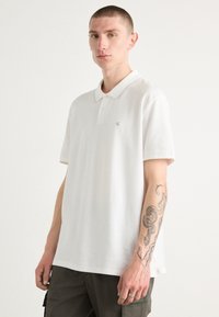 Hvitt polo skjorte med krage, korte ermer og subtilt logo på brystet. Stoffet virker glatt, med en rett skjæring.