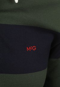 Gebreide trui met groene en marineblauwe horizontale strepen; heeft een rode geborduurde "McG"-logo aan de rechteronderzijde; ribbelstructuur.