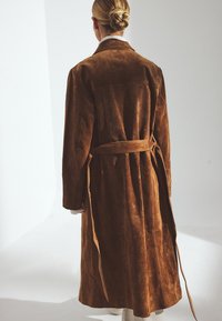 Brun suedetrenchcoat med bælte i taljen, lange ærmer og en blød tekstur. Har krave og store baglommer.