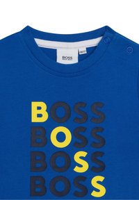 Modrá bavlněná trička s kulatým výstřihem a dvěma patentkami na ramenech. Výrazný nápis "BOSS" v žluté a tmavě šedé barvě uspořádaný vertikálně.