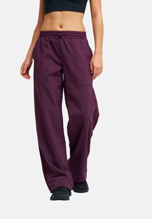 Femme portant un pantalon ample violet foncé avec de grandes poches à l'avant et des baskets noires, visible de la moitié du torse jusqu'aux pieds sur un fond uni.