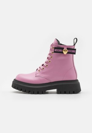 Stivale alla caviglia in pelle rosa con suola spessa nera, occhielli dorati e cinturino nero con un accento a forma di cuore dorato e il testo "MOSCHINO".