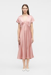 Γυναίκα που στέκεται φορώντας ένα μεταξένιο, ανοιχτό ροζ, μίντι φόρεμα με V-λαιμόκοψη και κοντά μανίκια, με λευκά μυτερά τακούνια, σε απλό λευκό φόντο.