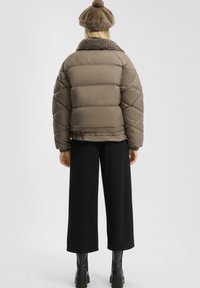 Kobieta ubrana w brązową kurtkę puchową z kołnierzem z polaru, pasującą brązową beretkę, czarne szerokie spodnie oraz czarne masywne botki, stojąca tyłem.