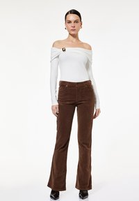 Weißes Off-Shoulder-Oberteil mit drapierten Ärmeln und goldenen Akzenten; kombiniert mit braunen Cordflared-Hosen. Glatter Stoff, maßgeschneiderte Passform.