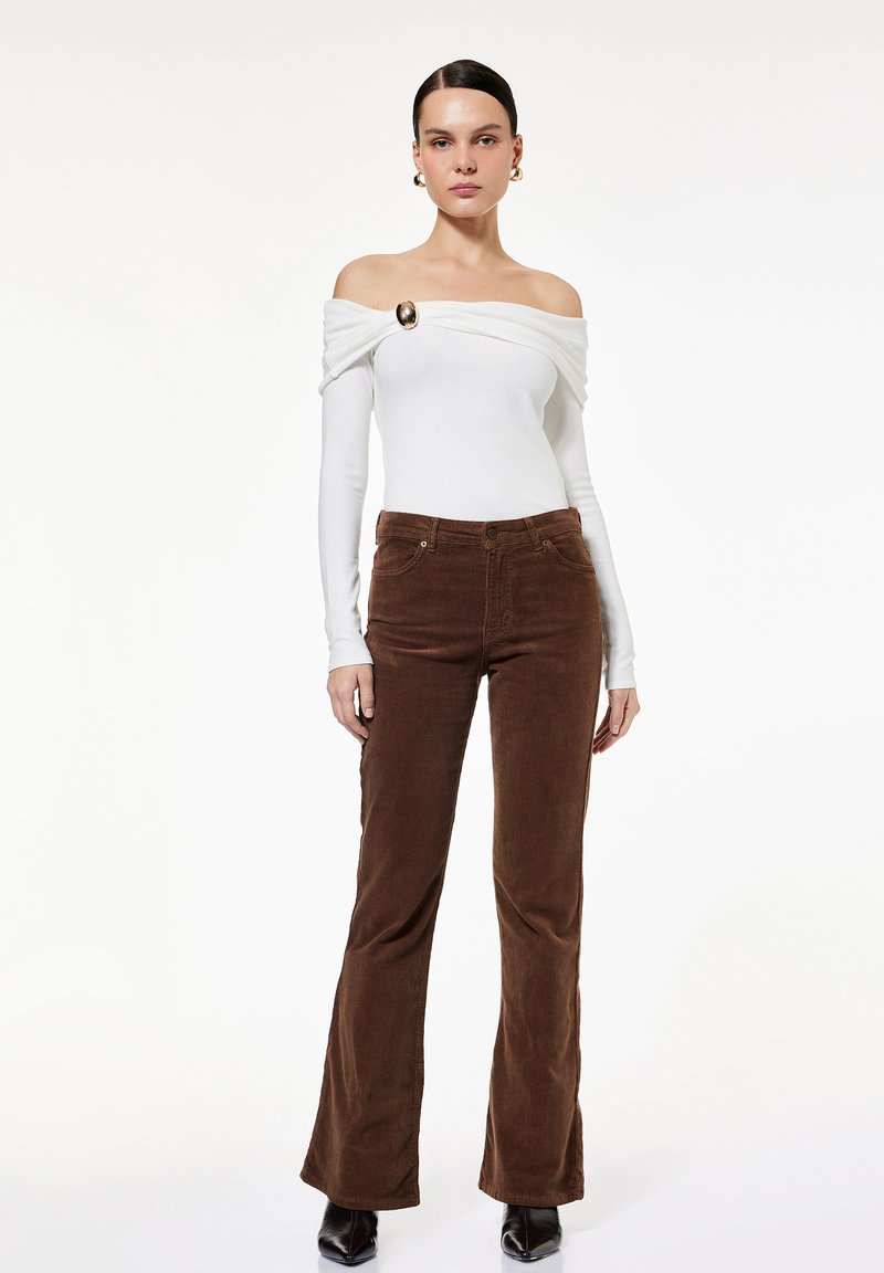 Weißes Off-Shoulder-Oberteil mit drapierten Ärmeln und goldenen Akzenten; kombiniert mit braunen Cordflared-Hosen. Glatter Stoff, maßgeschneiderte Passform.