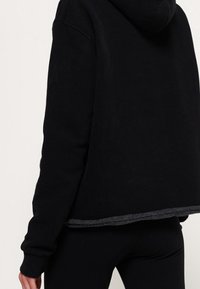 Svart huvtröja med avslappnad passform, som har en mjuk textur och en kontrasterande grå fåll. Enkel design utan synliga logotyper.