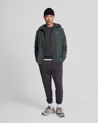 Veste légère verte avec capuche, sweat-shirt gris, pantalon de survêtement gris foncé et baskets. Détails notables incluent un logo jaune et un tissu texturé.