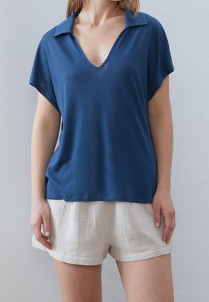 Femme portant un haut bleu ample à manches courtes en V et un short blanc texturé, debout devant un fond uni.