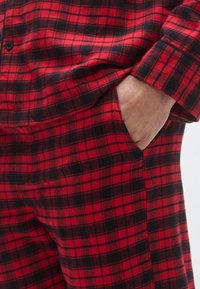 Tkanina flanelowa w czerwono-czarną kratę z kieszenią. Koszula ma zapięcie na guziki i mankiety, podkreślając przytulną teksturę.