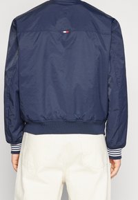 Navy blue bomber jacket with elastic cuffs and hem. Features a small red, white, and blue logo on the back. Smooth, lightweight material.