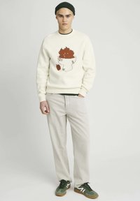 Sweat-shirt crème avec un graphisme de bol de fruits en marron, associé à un pantalon clair et des baskets vertes. Bonnet en tricot sur la tête.