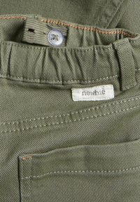 Pantalons vert olive en tissu texturé, avec une fermeture à bouton, une étiquette indiquant "newbie" et des détails de couture visibles.