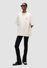 T-shirt crema oversize con logo ricamato, abbinato a leggings neri con fondo svasato e scarpe nere lucide. Gli accessori includono occhiali da sole neri piccoli.