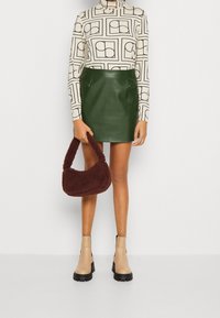 Mini gonna in pelle verde, top a maniche lunghe con motivo, borsa in pelle marrone a trama, e stivaletti beige con suola nera.