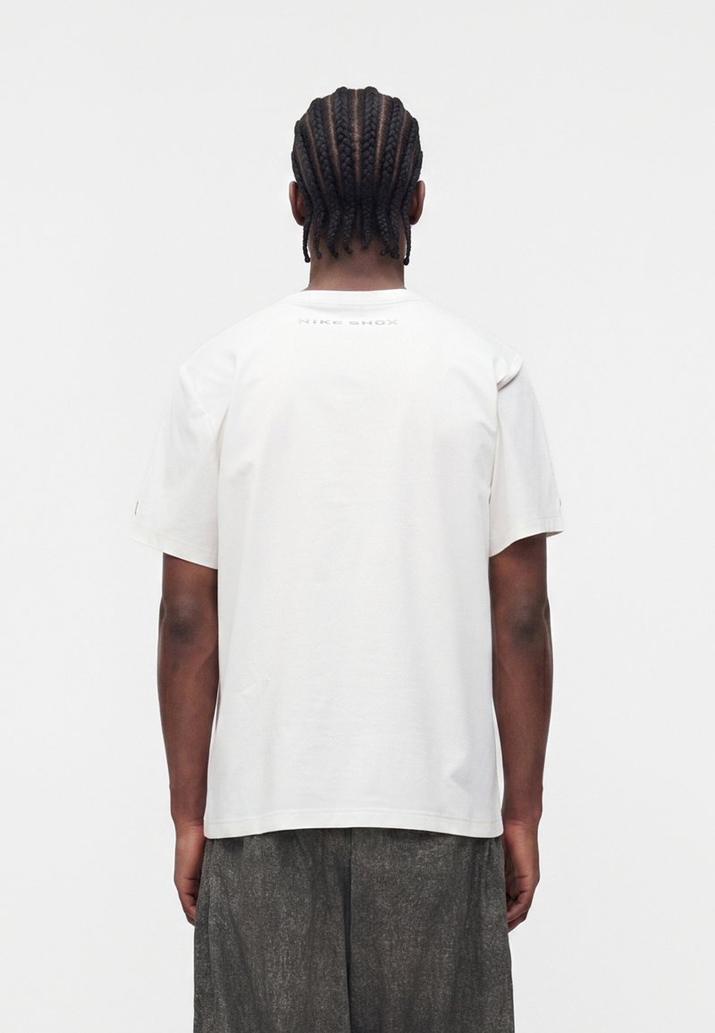 Man with braided hair wearing a plain white Nike Shox t-shirt and dark gray shorts, standing against a plain white background.