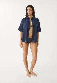 Camicia azzurra scuro con bottoni in tessuto leggero, maniche corte e finitura testurizzata, indossata sopra un bikini a pois.
