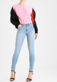 Jeans ajustados de color azul claro combinados con un suéter rosa de lana. La modelo lleva una chaqueta bomber negra con forro naranja y sandalias de tacón negras.